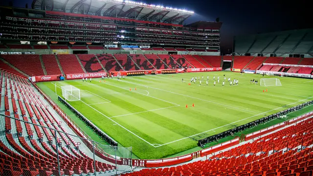 Interior del Estadio Caliente de Tijuana de noche, con las gradas rojas vacías y jugadores entrenando en la cancha